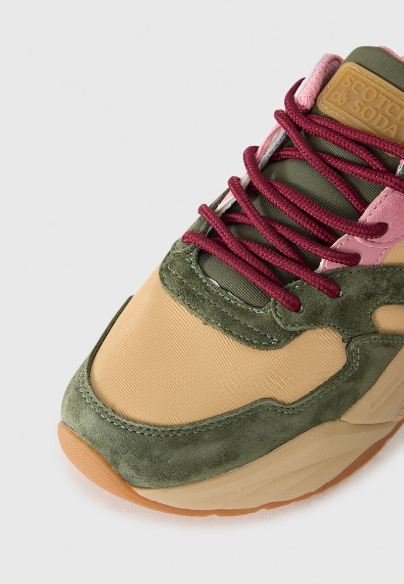Sneaker con una combinazione di suede beige e verde e tessuto, con lacci bordeaux e una suola in gomma testurizzata. Patch logo rettangolare ben visibile.