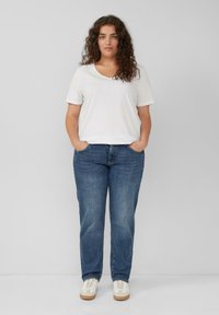 Biała koszulka z krótkim rękawem i dekoltem w szpic, połączona z niebieskimi dżinsami prostymi oraz białymi trampkami z beżowymi podeszwami. Model stoi z rękami w kieszeniach.