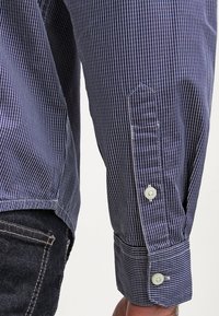 Gros plan d'une personne portant une chemise à manches longues rayée bleu et blanc avec des poignets boutonnés et un jean sombre.