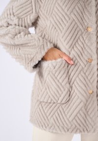 Lichtgrijze nepbontjas met een structuur in een visgraatpatroon, twee gouden knopen en een zijzak. Zacht en pluizig materiaal.