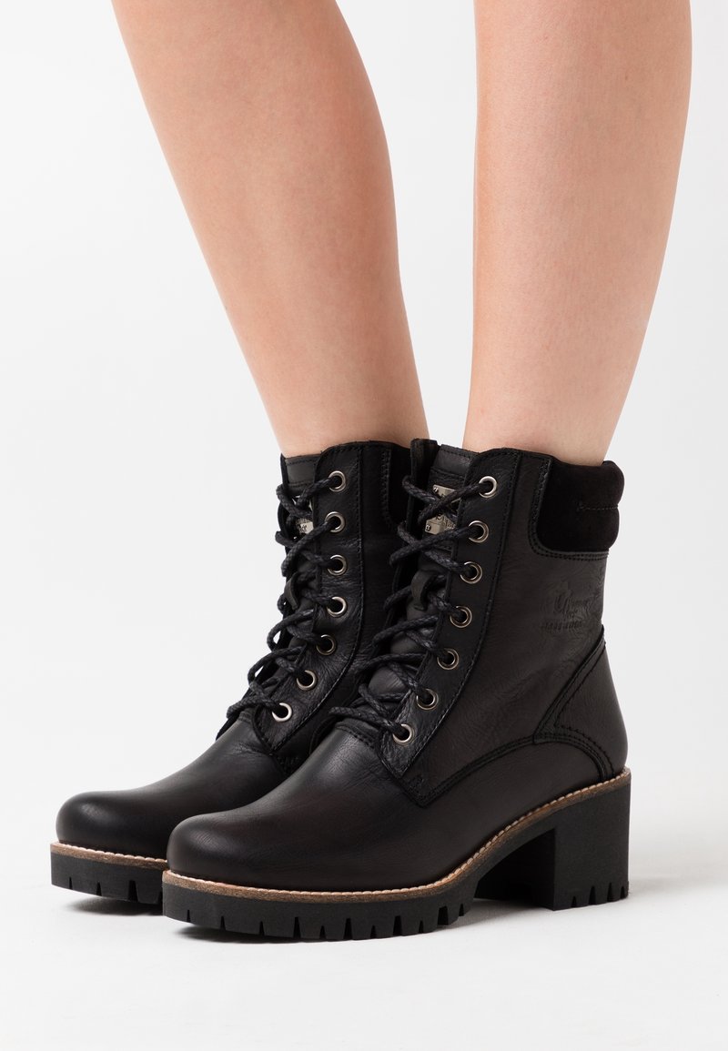 Bottines noires en cuir à lacets avec talons carrés, portées sur des jambes nues sur fond blanc.