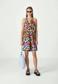 Mulher vestindo um vestido halter floral colorido, sandálias plataforma pretas, óculos de sol e segurando uma bolsa de mão azul clara em um fundo simples.