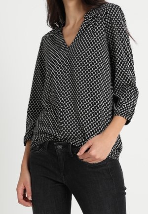 Nainen mustassa bleiserissä, jossa on valkoiset timanttikuvioinnit, ja mustissa farkuissa, toinen käsi taskussa, yksiväristä taustaa vasten.