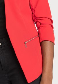 Blazer rojo hecho a medida de tela suave con un detalle de bolsillo con cremallera. Presenta mangas fruncidas y una silueta elegante.