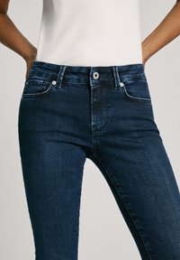 Jeans de mezclilla azul oscuro con un diseño ajustado, cintura de altura media, bolsillos frontales y detalles de costura sutiles. Textura suave y cierre clásico con botón.