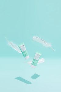 Två mintgröna rör med solskyddskräm, med vita etiketter och svart text, som sväva över mjuka vita fjädrar mot en ljus bakgrund.