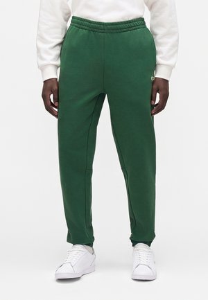 Mørkhudet person iført grønne joggingbukser, hvid sweatshirt og hvide Lacoste-sneakers står mod en ensfarvet baggrund.