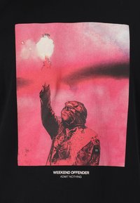T-shirt noire avec un imprimé graphique représentant une figure portant un masque et des lunettes, levant une main sur un fond rose, avec du texte en dessous.
