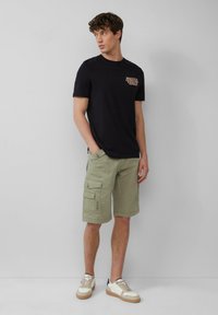 Zwarte katoenen t-shirt met grafisch ontwerp op de borst, gecombineerd met lichtgroene cargo shorts met meerdere zakken, en beige sneakers met witte accenten.