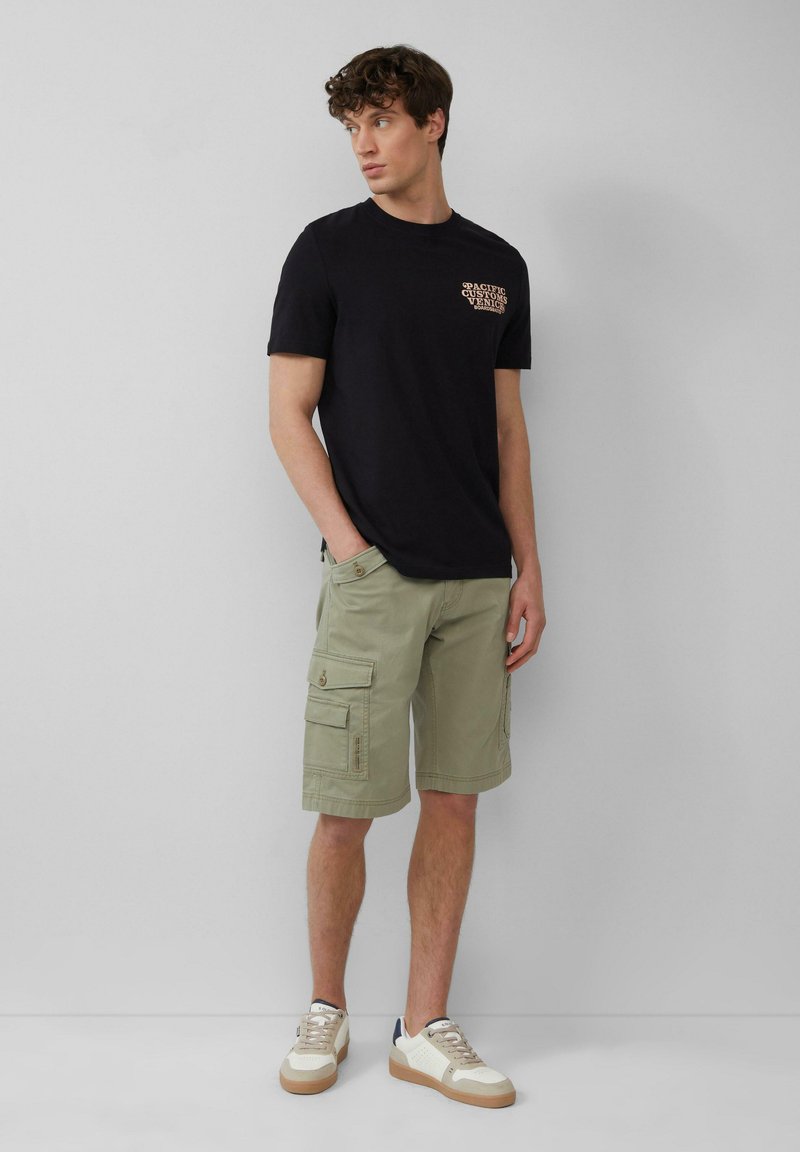 Zwarte katoenen t-shirt met grafisch ontwerp op de borst, gecombineerd met lichtgroene cargo shorts met meerdere zakken, en beige sneakers met witte accenten.