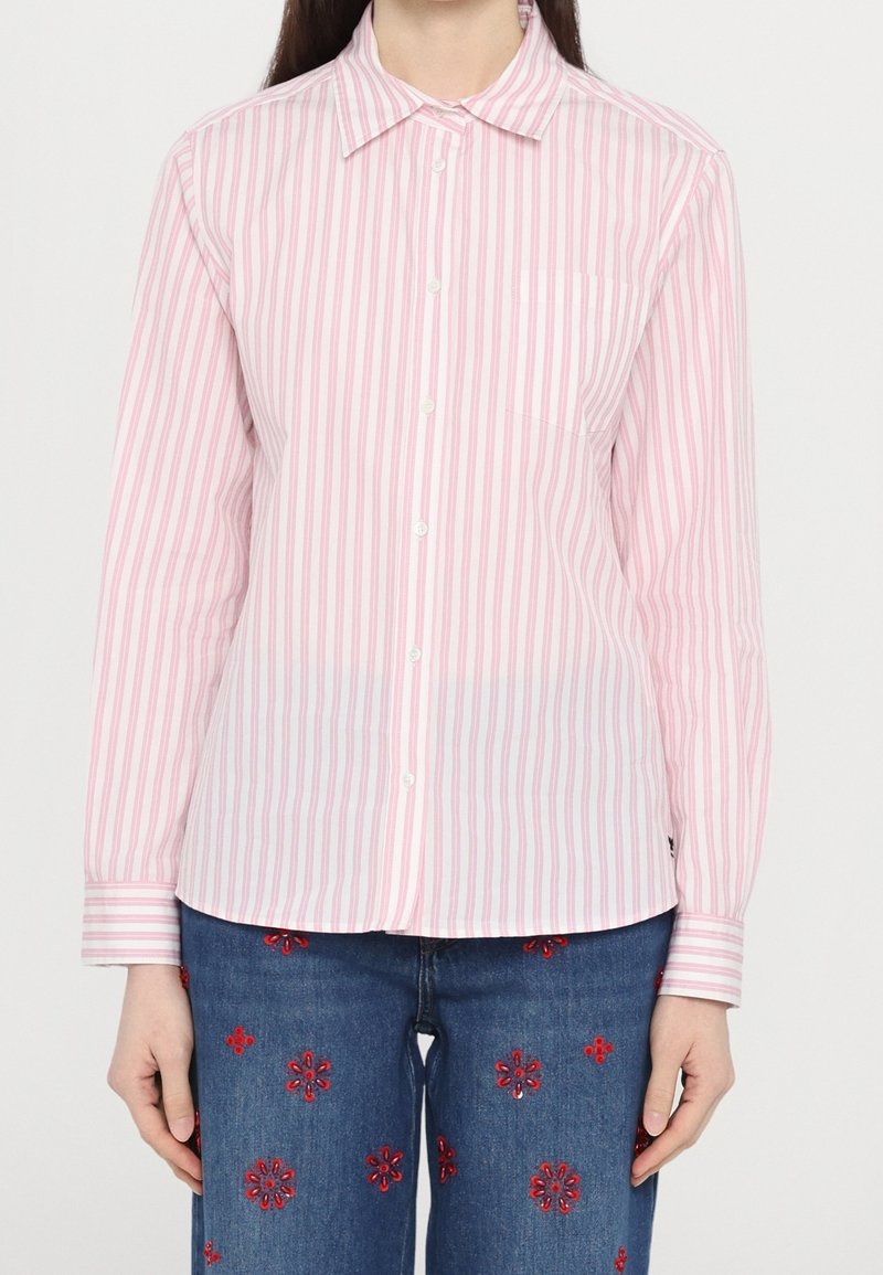Femme portant une chemise boutonnée à rayures verticales rose et blanche avec un jean bleu brodé de motifs floraux rouges.