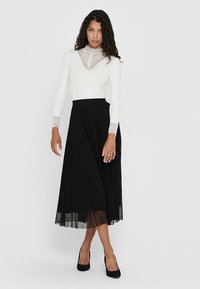 Jupe plissée noire longue associée à un haut à manches longues blanc avec un col en dentelle et des manchons. La tenue est complétée par des chaussures noires.