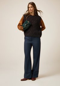 Gilet noir en tricot avec un motif texturé, chemise à manches longues marron en dessous, pantalon large bleu marine et sac à main vert. Des chaussures marron complètent le look.