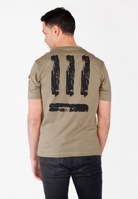 Camiseta de manga corta color verde oliva con un gran diseño de pinceladas negras en la parte posterior. Fabricada en algodón suave.