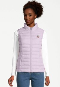 Chaleco acolchado de color púrpura claro con cuello alto, cierre de cremallera y costuras horizontales. Presenta un pequeño logo en el pecho. Lleva puesto sobre una camiseta de manga larga blanca.