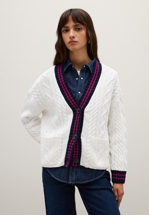 White cable knit cardigan with dark navy and fuchsia striped vee collar and cuffs. Two front pockets and button closure. Worn over denim shirt.