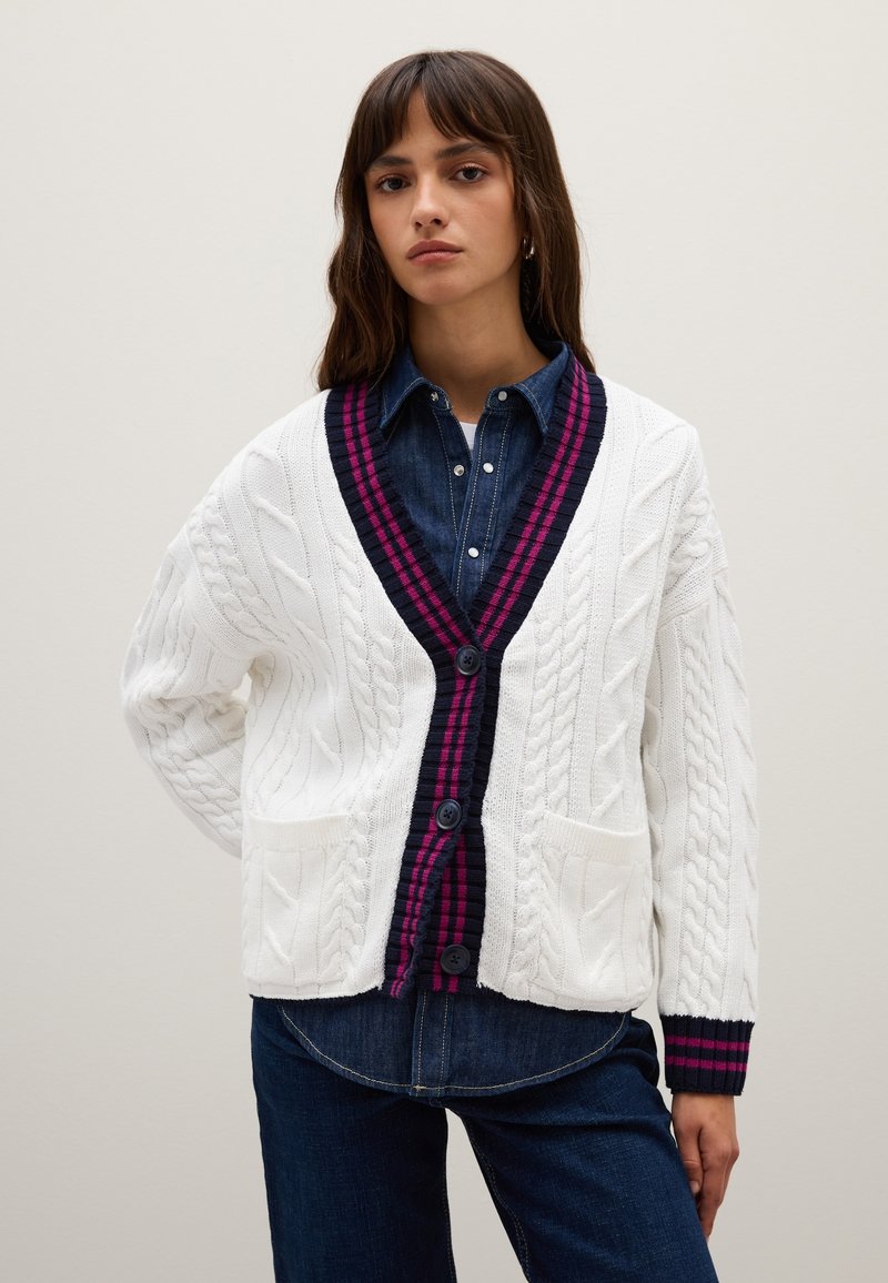 Weißer Kabelstrick-Cardigan mit dunkelblauem und fuchsiafarbenem gestreiften V-Kragen und Bündchen. Zwei aufgesetzte Taschen und Knopfverschluss. Über einem Jeanshemd getragen.