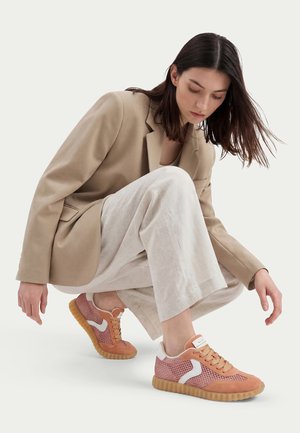 Femme accroupie portant un blazer beige, un pantalon large clair et des baskets en maille rose avec des accents blancs et des semelles gomme sur un fond uni.