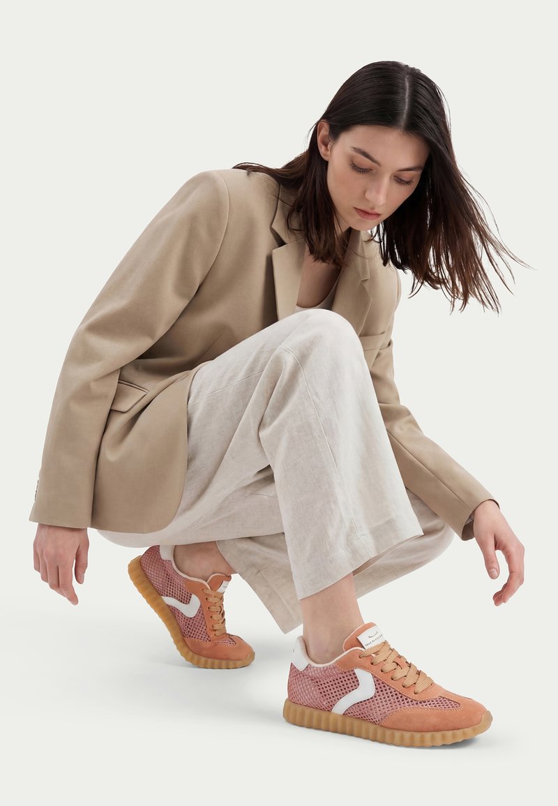 Frau hockt, trägt einen beigen Blazer, helle weite Hosen und rosa Mesh-Sneaker mit weißen Akzenten und braunen Sohlen vor einem einfarbigen Hintergrund.