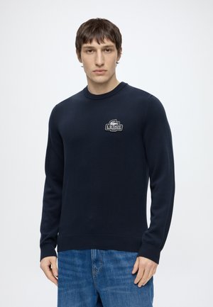 Homme portant un pull Lacoste bleu marine avec logo sur la poitrine et un jean bleu, debout devant un fond blanc uni.