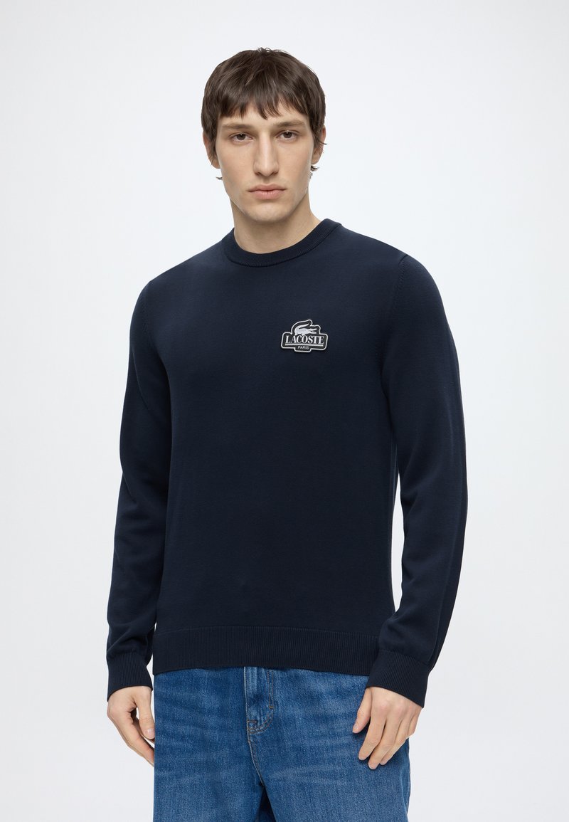Homme portant un pull Lacoste bleu marine avec logo sur la poitrine et un jean bleu, debout devant un fond blanc uni.