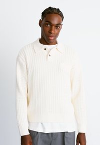 Suéter de punto color crema con un patrón texturizado, que presenta un cuello con solapa y tres botones. Incluye un pequeño bolsillo en el pecho.
