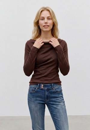 Femme aux longs cheveux blonds portant un chemisier marron foncé à manches longues et un jean bleu, tenant l'encolure de son chemisier avec les deux mains.