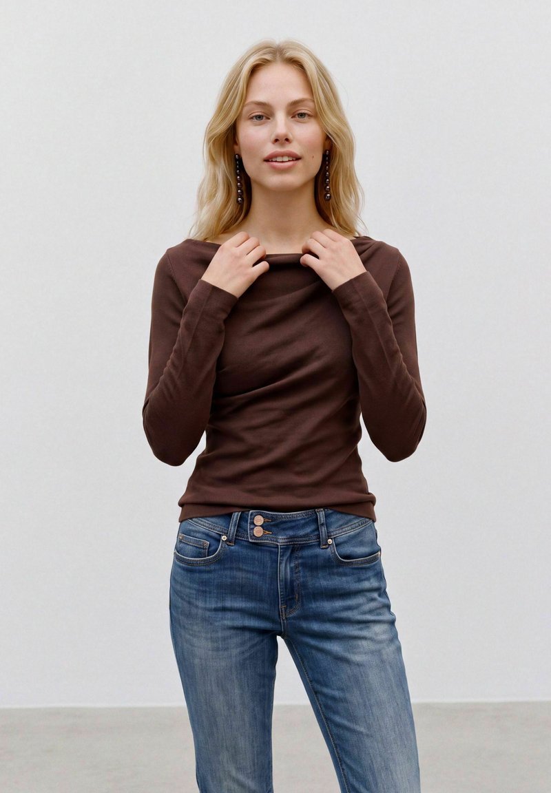 Femme aux longs cheveux blonds portant un chemisier marron foncé à manches longues et un jean bleu, tenant l'encolure de son chemisier avec les deux mains.
