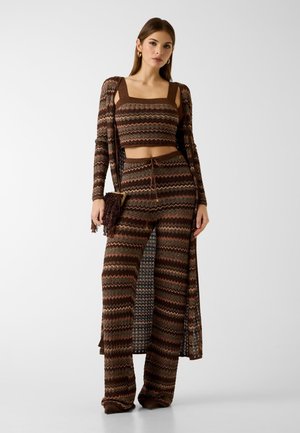 Donna che indossa un top corto marrone a zigzag lavorato a maglia, pantaloni a gamba larga abbinati, un cardigan lungo e tiene una borsa marrone con frange, in piedi su sfondo bianco.