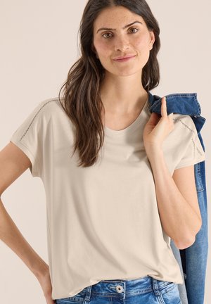 Donna con capelli lunghi e castani che indossa una maglietta beige a maniche corte, con una giacca di jeans blu appoggiata sulla spalla, che sorride leggermente.