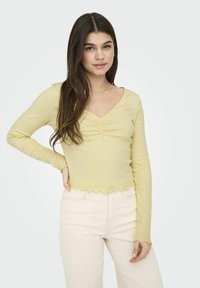 Langdurige gele top met een V-hals, voorzien van een geraffineerd midden en een scalloped zoom. Gecombineerd met lichtgekleurde broek.