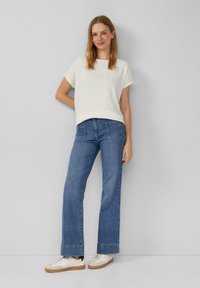 Witte, textuurgegeven top met korte mouwen in combinatie met blauwe wijde jeans. Witte sneakers met tan kleurige zolen. Eenvoudige, casual outfit.