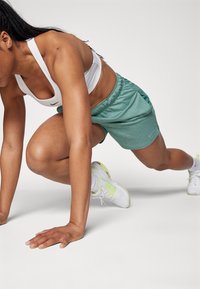 Sujetando una posición de cuclillas, viste un sujetador deportivo de Nike en blanco, pantalones cortos de sudor verdes y zapatillas blancas con acentos amarillos.
