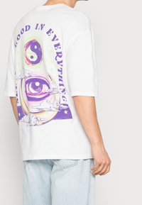Camiseta de algodón blanca que presenta un gráfico vibrante en la parte posterior con un ojo, el símbolo del yin-yang, nubes y texto en púrpura sobre un fondo claro.