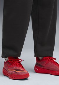 Røde atletiske sko med struktureret stof, kontrasterende orange stribe og tonale snørebånd. Parret med sorte sweatpants, der viser en afslappet pasform.