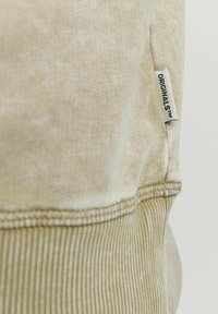 Sudadera beige con tela texturizada, dobladillo acanalado y costuras visibles. Presenta una pequeña etiqueta blanca con la inscripción "ORIGINALS™."