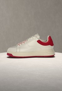 Λευκό sneaker χαμηλού ύψους με κόκκινες λεπτομέρειες στη φτέρνα, τα κορδόνια και την σόλα, που εμφανίζεται σε φωτεινή επιφάνεια με απλή γκρι φόντο.