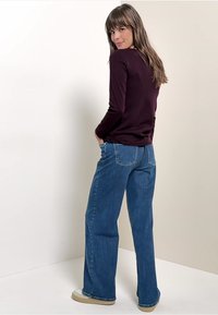 Dunkellila langärmliges Oberteil, kombiniert mit blauen hoch taillierten weitem Jeans. Die Jeans verfügen über Gesäßtaschen und einen lässigen Schnitt, ergänzt durch leichte Schuhe.