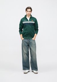 Zelená bunda na zips s bielymi a námorníckymi pruhmi, kombinovaná s širokými svetlomodrými džínsami a bielymi teniskami, stojaca na jednofarebnom pozadí.