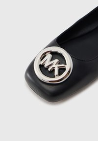 Μαύρο δερμάτινο παπούτσι με φλατ σόλα, που διαθέτει ένα εξέχον ασημί κυκλικό λογότυπο με τα γράμματα "MK", στρογγυλεμένη μύτη και ομαλή υφ�ή.