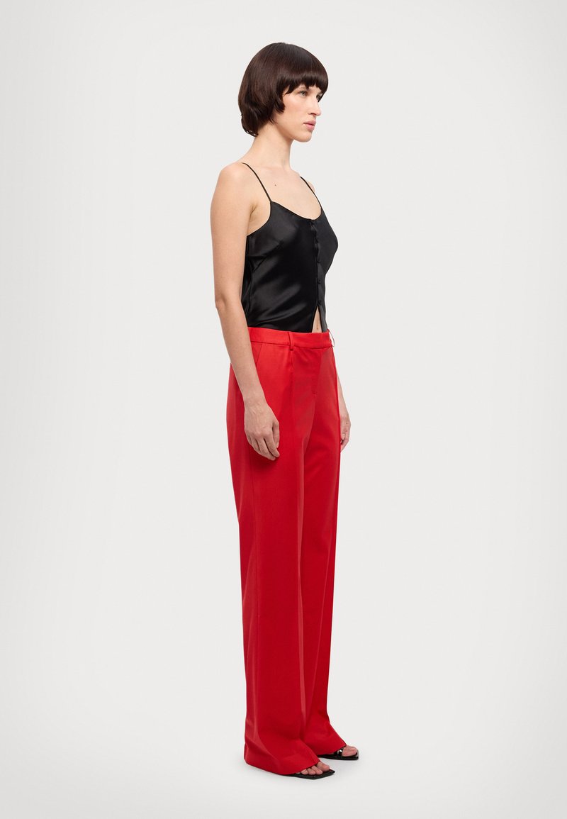 Black satin camisole with button detail, paired with high-waisted, tailored red trousers. Model wears black open-toe sandals.