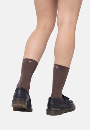 Braune kniehohe Socken mit geripptem oberen Rand, versehen mit einem kleinen roten und weißen Akzent nahe dem Bündchen, kombiniert mit schwarzen Lederschuhen.