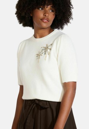 Pull en tricot blanc à manches courtes, avec un brodé de palmier en sequins à l'avant. Associé à une jupe marron foncé.