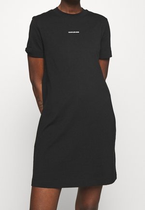 Schwarzes T-Shirt-Kleid mit kurzen Ärmeln und kleinem weißen "Calvin Klein Jeans"-Logo auf der Brust, getragen von einer Person mit hinter dem Rücken verschränkten Händen.