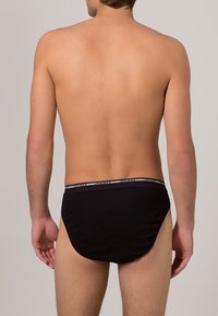 Schwarze Baumwollslips mit einem weichen elastischen Bund, auf dem der Markenname "JOCKEY" in Grau zu sehen ist, von hinten betrachtet. Glatte Textur.