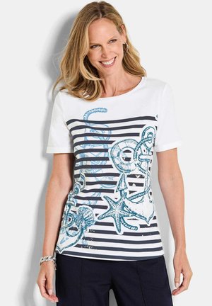 Lächelnde Frau mit blonden Haaren trägt ein weißes Kurzarmoberteil mit nautischen schwarzen Streifen und blauen, meeresinspirierten Designs, kombiniert mit dunkler Hose.