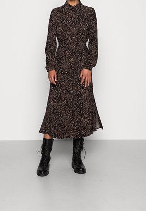 Vrouw die een donkeraanimalprint midi-jurk met lange mouwen en knopen draagt, gecombineerd met zwarte veterlaarzen, tegen een effen achtergrond.