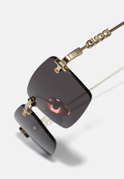 Ochelari de soare pătrați cu lentile întunecate, accente din metal aurit și balamale laterale elegante. Dispune de un logo proeminent pe bratul ochelarilor.