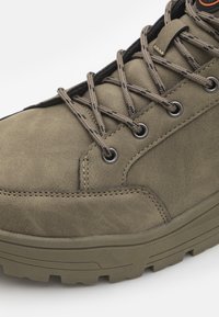 Bota de cuero verde oliva con superficie texturizada, puntera redonda, ojales de metal y cordones encerados; suela de goma gruesa con surcos profundos para tracción.