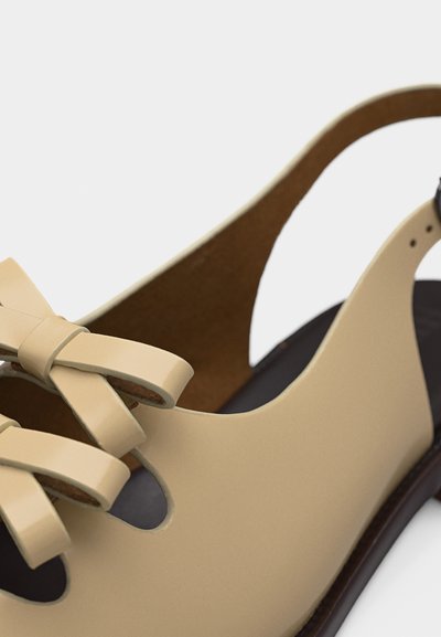 Μπεζ δερμάτινο παπούτσι slingback με ένα διακοσμητικό δερμάτινο φιόγκο στο μπροστινό μέρος και σκούρα καφέ σόλα.